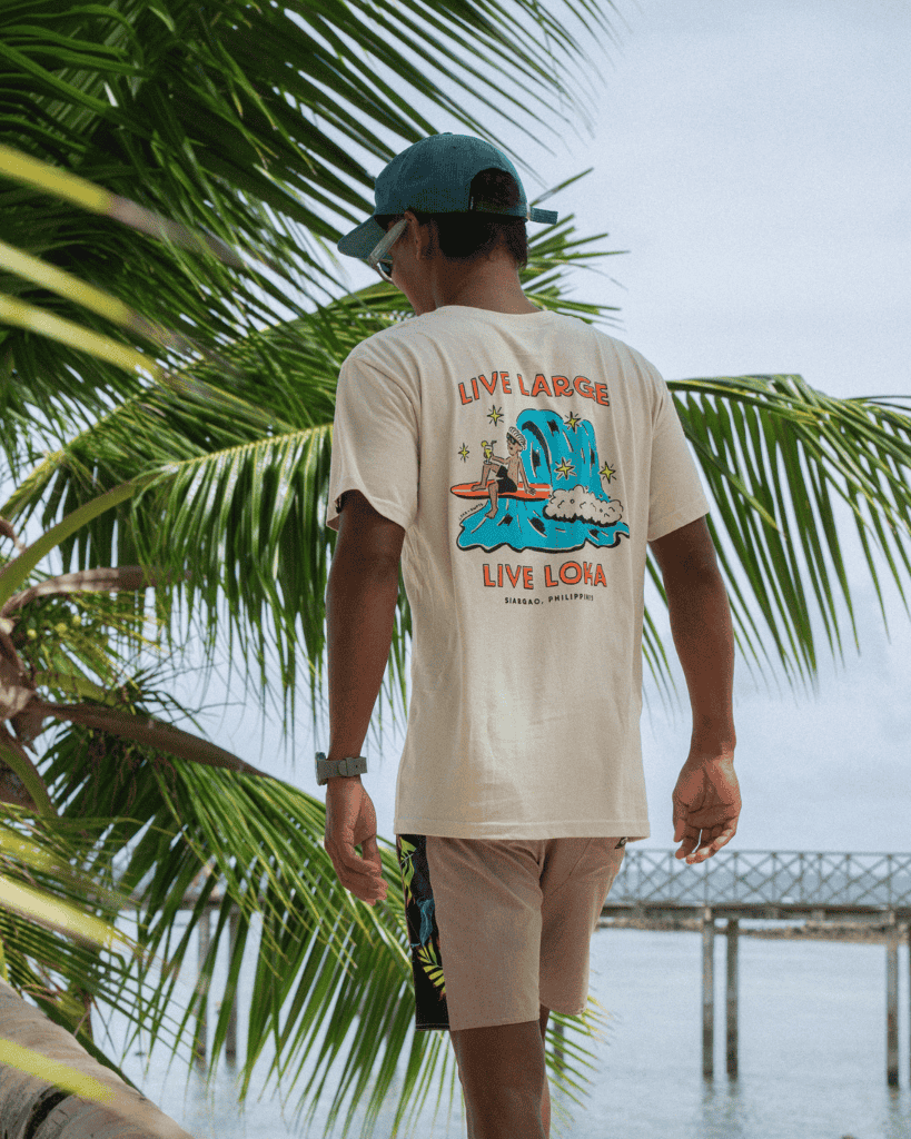 Person wearing a beige t-shirt with graphics and text, standing under palm leaves by a body of water.