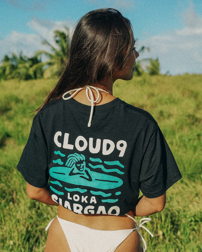 A person wearing a black crop top with 'CLOUD9 LOKA SIARGAO' text and a graphic on the front, paired with white bottoms, standing in a field.