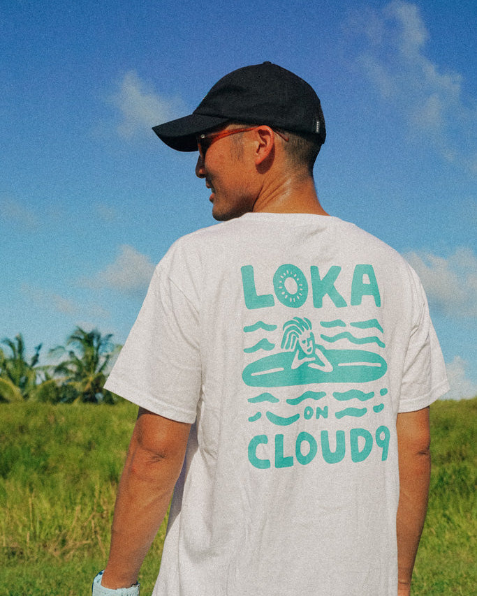 A person wearing a white crew neck t-shirt with text and graphic design facing away from the camera, standing in an outdoor setting.