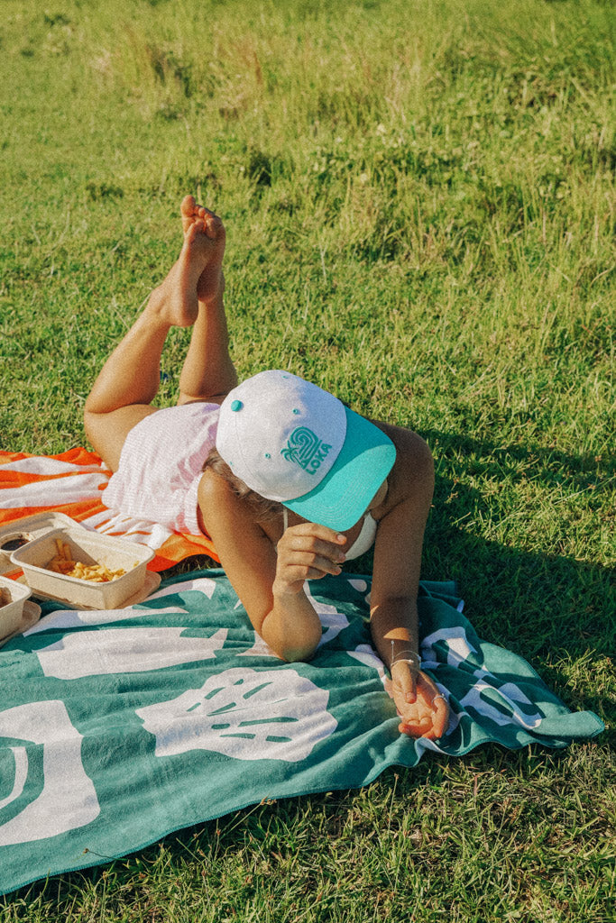 A person lying on a blanket wearing a white 'Swell Dad Cap' from the On Cloud 9 Collection.