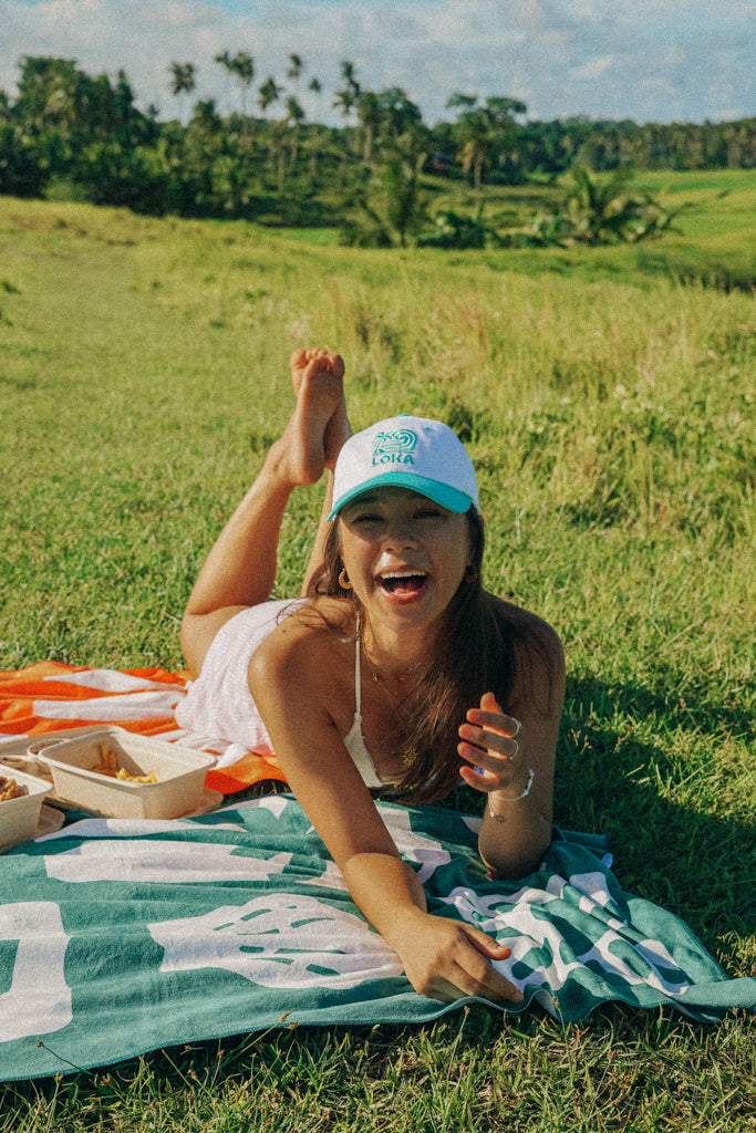 A person lying on a blanket wearing a white 'Swell Dad Cap' from the On Cloud 9 Collection.